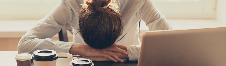 Exhausted businesswoman slumped over desk with laptop and coffee cups.