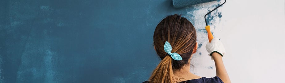 Girl uses roller to paint white wall teal blue.